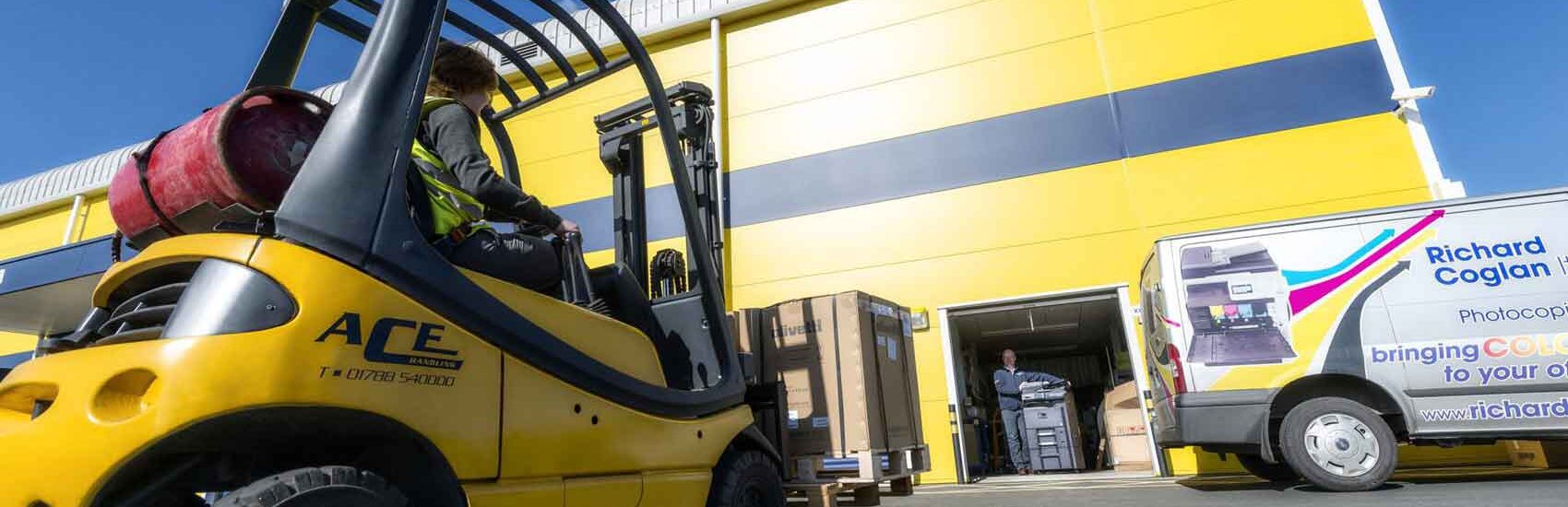 Forklift unloading boxes outside a yellow storage facility with delivery van nearby