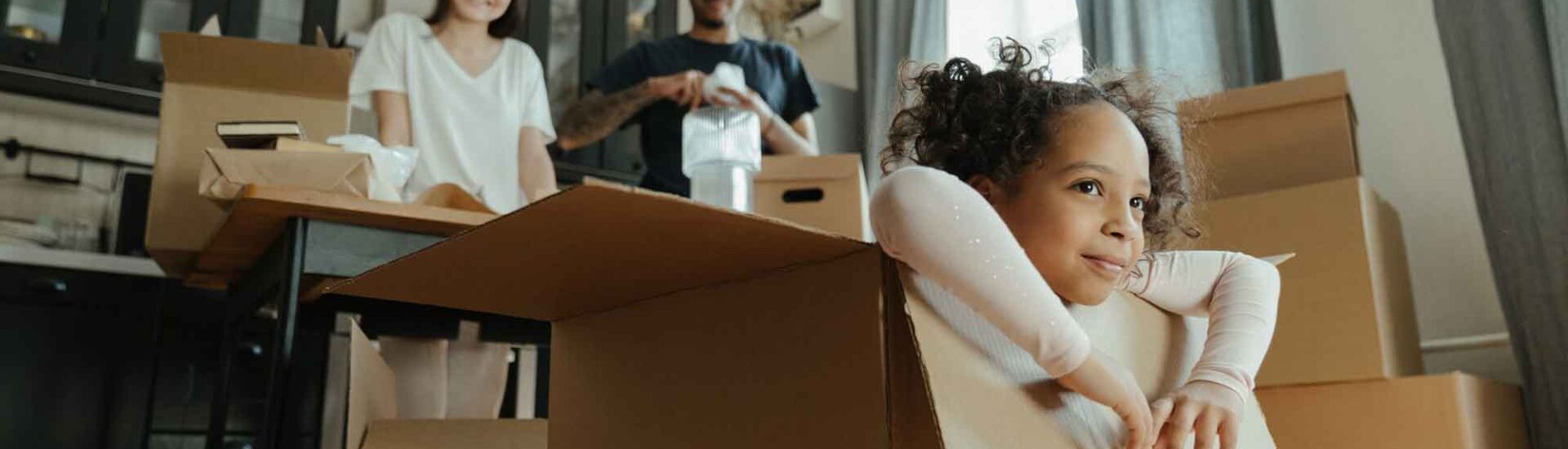 Smiling child playing inside a cardboard box while parents pack up their home in the background