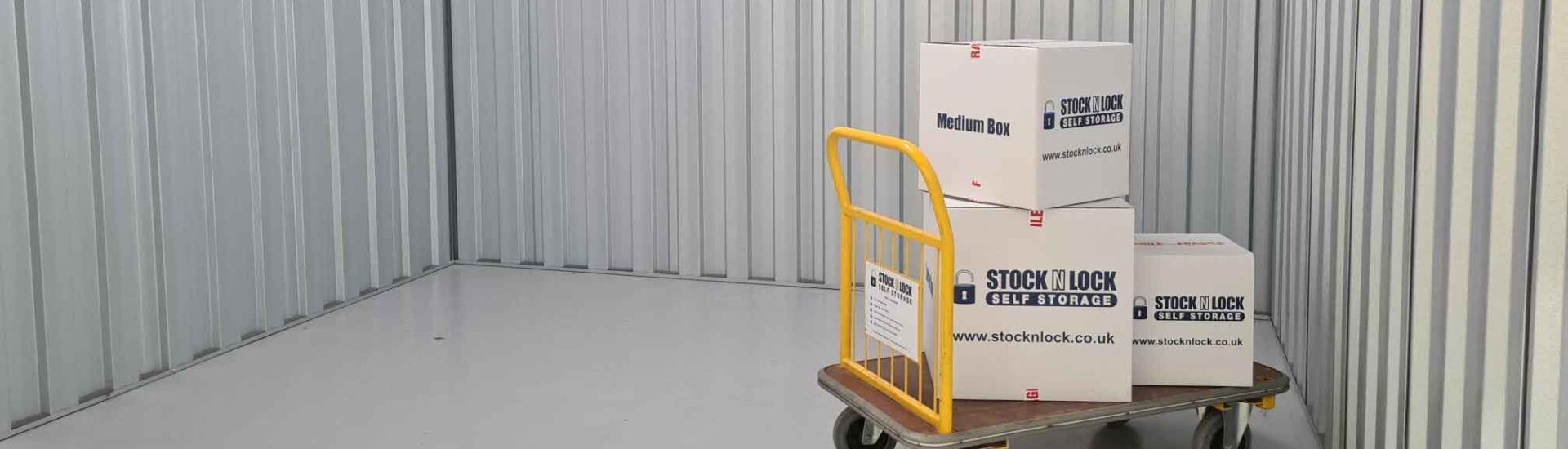 Stock N Lock branded boxes on a yellow trolley inside a clean, empty self-storage unit with metal walls
